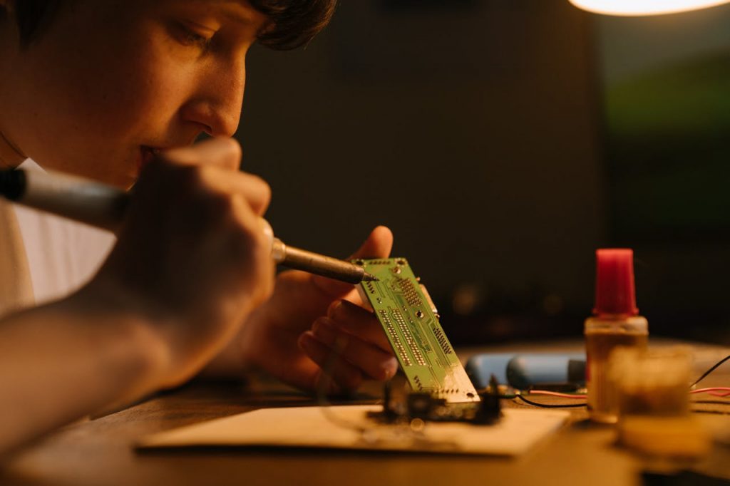 Focused young technician soldering a printed circuit board indoors, showcasing DIY electronics skills.